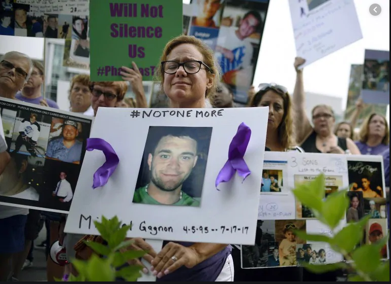 A mourning woman at an Opioid Crisis rally holds a poster featuring a young man she lost to the crisis. 

The poster uses the social media tag "#NotOneMore".